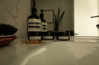 One And Only and Aesop face products and hand soap, with a cherry red La Bonne Brosse hairbrush, arranged on a white bathroom counter next to a mirror and gold jewelry.